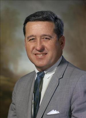 A head and shoulders portrait of a white man with short hair, a gray suit jacket, and a dark tie. He has a white hanky peaking out of his chest pocket. 